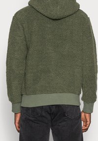 Giacca in pile testurizzata verde oliva con cappuccio indossata da una persona di spalle, abbinata a jeans in denim neri.