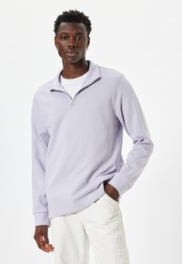 Suéter de media cremallera en color púrpura claro, hecho de una tela suave, con mangas largas y un corte holgado, combinado con pantalones de color claro.