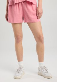 Pantalones cortos de lino rosa con cintura elástica y cordón. Combinados con zapatillas blancas y calcetines. Textura suave, por encima de la rodilla.