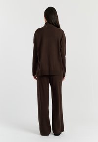 Pull en tricot marron foncé avec un col montant, des épaules tombantes et des fentes sur les côtés, associé à un pantalon large assorti. Tissu texturé et simple.
