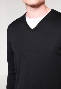 Maglione nero a V con maniche lunghe, realizzato in tessuto a maglia testurizzato. Presenta una camicia bianca con colletto rotondo sotto.