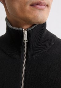 Schwarzer Strickpullover mit hohem Kragen, grauem geripptem Innenkragen, silbernem Frontreißverschluss und strukturiertem Wollmaterial.
