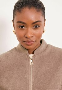 Chaqueta de lana beige con cremallera, cuello alto y cremallera dorada. La modelo lleva pendientes de aro dorados y tiene el cabello liso y oscuro. Tejido sencillo con textura.