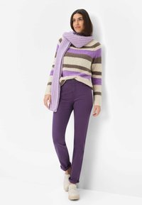 Maglione a righe viola e beige, sciarpa lavorata a maglia e pantaloni viola, abbinati a calzature di colore chiaro, su sfondo semplice.