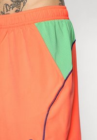 Pomarańczowe szorty sportowe z zielonym siateczkowym panelem bocznym, elastycznym paskiem w talii i kontrastowym niebieskim szwem wzdłuż szwów.
