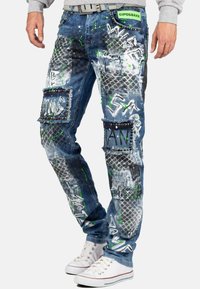 Jeans en denim avec une finition déchirée, comportant des patchs, des imprimés de style graffiti, des accents verts et des embellissements en clous argentés.