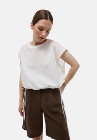 Haut blanc ample en tissu léger, à manches courtes et avec un ourlet resserré à l'aide de liens réglables. Associé à un short rayé marron.