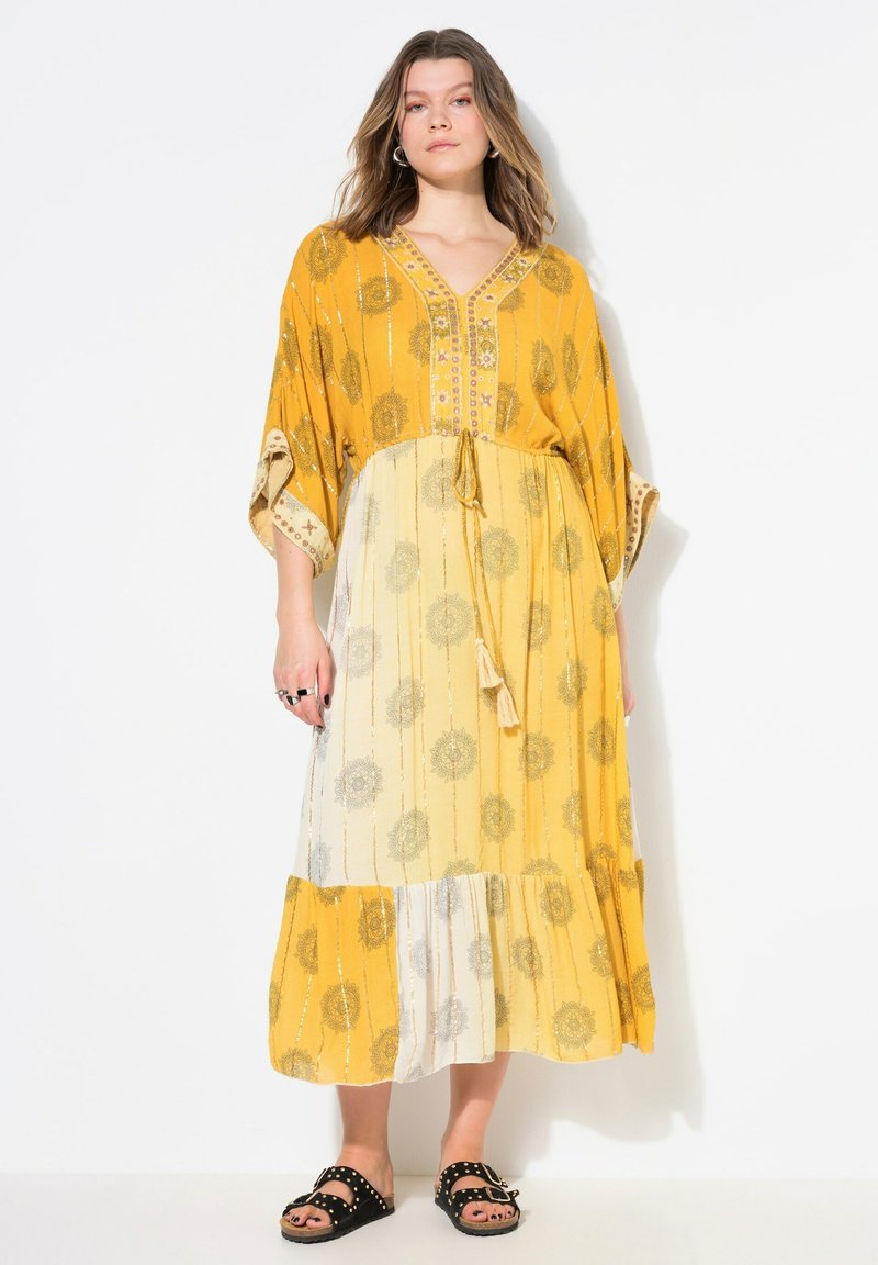 Abito maxi giallo senape con motivi floreali, cintura in vita e orlo a strati. Tessuto leggero con vestibilità comoda e maniche a pipistrello.