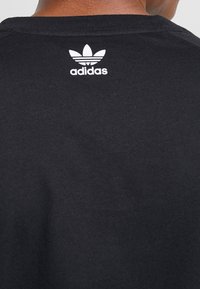 Baksidan av en persons nacke som bär en svart Adidas-tröja med den vita Adidas-logotypen och trefoil-symbolen centrerad under halsringningen.