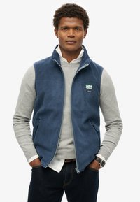 Granatowa kamizelka poliestrowa z zamkiem z przodu, dwoma bocznymi kieszeniami i naszywką z logo. Noszona na jasnoszarym, długim rękawie i ciemnych dżinsach.
