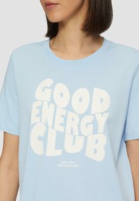 Lichtblauwe katoenen t-shirt met korte mouwen, met vetgedrukte witte tekst "GOOD ENERGY CLUB" en kleinere tekst "JUST HAPPY VIBES OVER HERE."