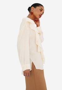 Blusa crema con dettagli arricciati sul davanti, maniche lunghe e una vestibilità leggermente ampia. Abbinata a pantaloni marroni, che mostrano un tessuto testurizzato.
