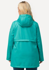 Kalkblå vattentät jacka med huva, med en åtsittande baksida och en horisontell söm, samt en avslappnad siluett. Slät, glänsande yta.