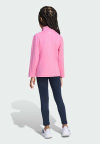 Fleece bunda v jasně růžové barvě s vysokým límcem, v kombinaci s námořnickými legínami a bílými sportovními botami. Texturovaná tkanina, volný střih.