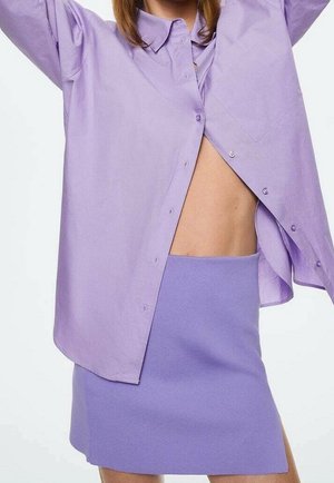 Vrouw draagt een lichtpaarse oversized blouse, opengeknoopt om de blote buik te tonen, en een nauwsluitende paarse minirok met een zijsplit.