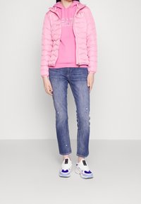 Světle růžová prošívaná péřová bunda, zkombinovaná s růžovou mikinou s kapucí, modrými džínami s širšími okraji a bílými teniskami s modrými detaily.