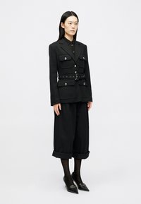 Musta muotoiltu takki, jossa on useita etutaskuja ja hopeanvärisiä nappia, yhdistettynä leveälahkeisiin mustiin housuihin ja kärkikengät.