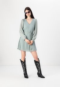 Lysegrønn teksturert kjole med V-hals, lange transparente ermer og belte i midjen. Brukt med svarte broderte cowboystøvler og solbriller.