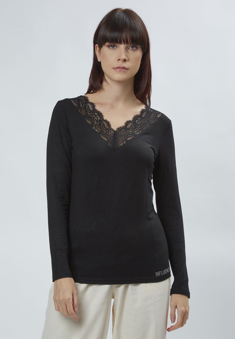 Schwarzes Langarmshirt mit einem Spitzen-V-Ausschnitt. Weicher Stoff, taillierte Form und das Wort "INFLUENCE" in einem metallischen Akzent am Saum.