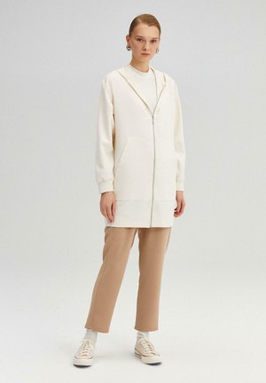 Sudadera con capucha cremosa con cremallera, bolsillos frontales, puños elásticos, combinada con pantalones beige ajustados y zapatillas blancas. Textura de tela suave.