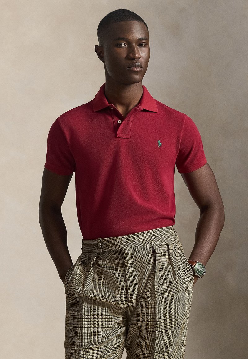 Άνδρας που φορά κόκκινο πουκάμισο  πόλο και μπεζ καρό πιετέ παντελόνι με τα χέρια στις τσέπες, μπροστά σε ουδέτερο φόντο.