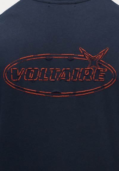 Námořnicky modrý textil zdobí výrazné vyšívané logo "VOLTAIRE" v oranžové barvě uvnitř oválu. Látka má jemnou texturu a známky opotřebení.