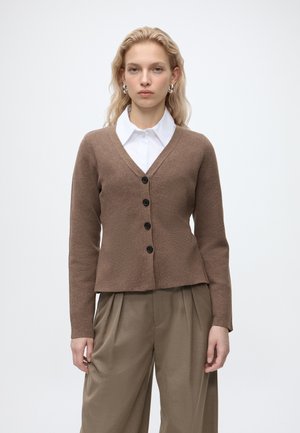 Femme aux cheveux blonds porte un cardigan marron boutonné sur une chemise blanche à col et un pantalon beige ample sur fond blanc uni.