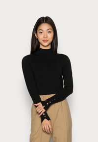 Schwarz langärmliges Rollkragenoberteil mit figurbetontem Design und geripptem Textur, mit Knopfdetails an den Bündchen. Kombination mit beigen hoch taillierten Hosen.