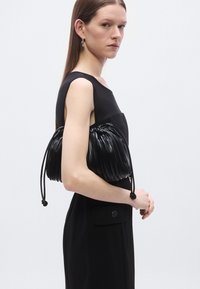 Svart veske i kunstlær med vertikale folder, snorstramming og rund form. Modellen har på seg et stilrent svart antrekk.