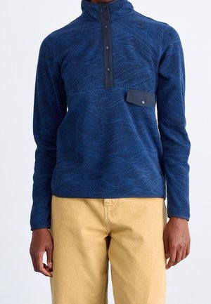 Personne portant un pull à motifs bleus avec des boutons-pression noirs et un pantalon beige devant un fond clair et uni.