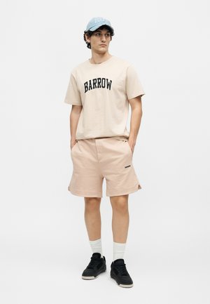 Joven con camiseta y pantalones cortos beige BARROW, zapatillas negras, calcetines blancos y gorra azul claro, de pie sobre fondo blanco.