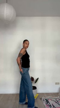 Žena s tetovažama u crnoj majici bez rukava i širokim trapericama stoji bočno u minimalističkoj sobi s biljkom i uzorkovanim tepihom.