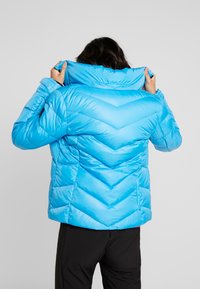 Veste matelassée bleu clair avec un design en chevrons et un col haut, présentant une texture lisse et brillante ainsi que des poignets élastiqués.