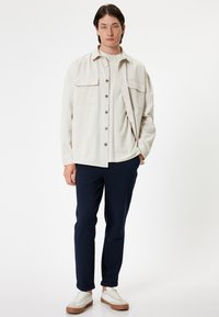 Lichtbeige jas met twee borstzakken, crèmekleurig overhemd, marineblauwe broek en witte sneakers met tan accenten. Glad materiaal en ontspannen pasvorm.