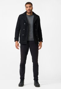 Schwarzer doppelt-knöpfiger Wollmantel aus strukturiertem Material, getragen über einem grauen Pullover, kombiniert mit schwarzen Hosen und Schnürstiefeln.