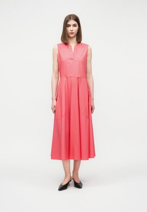 Femme debout portant une robe midi corail sans manches avec un décolleté en V et des chaussures noires à talons bas, sur un fond clair uni.