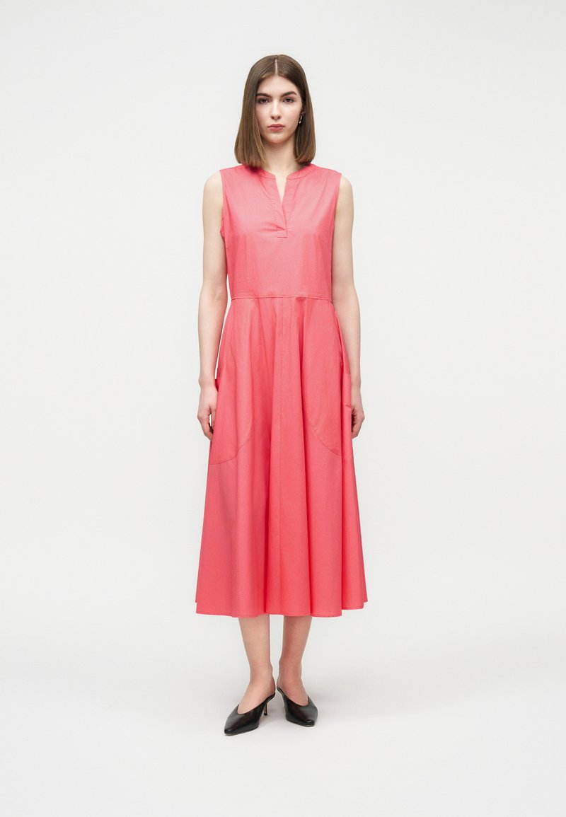 Femme debout portant une robe midi corail sans manches avec un décolleté en V et des chaussures noires à talons bas, sur un fond clair uni.