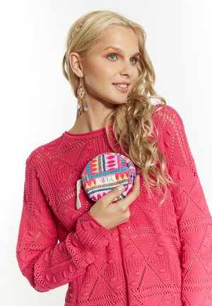 Maglione lavorato a maglia di un luminoso colore rosa con motivi intricati; tiene un sacchetto circolare multicolore con design geometrici e un'etichetta.