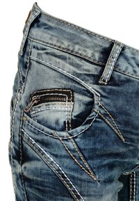 Gros plan d'un jean en denim bleu délavé montrant une poche avant avec une petite poche zippée pour pièces et des coutures détaillées blanches et marron.