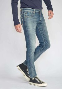 Jean en denim bleu avec une coupe slim, comporte cinq poches et une fermeture éclair. Porté avec des baskets montantes noires et une chemise à manches longues bleu marine.