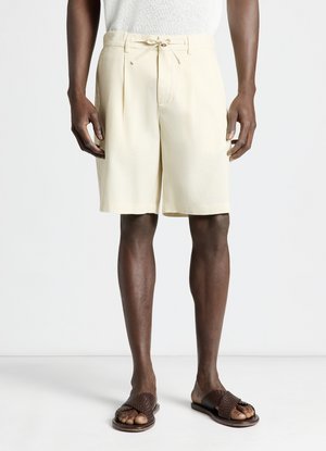 Homme portant un short crème sur mesure avec une taille à cordon et des sandales marron tissées à enfiler, debout sur un sol blanc.