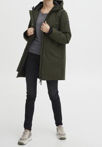 Khaki parka s kapucí, zipem a patentkami, dvěma předními kapsami, pletenými manžetami a drobnou nášivkou loga, spárovaná s tmavými džíny a teniskami.