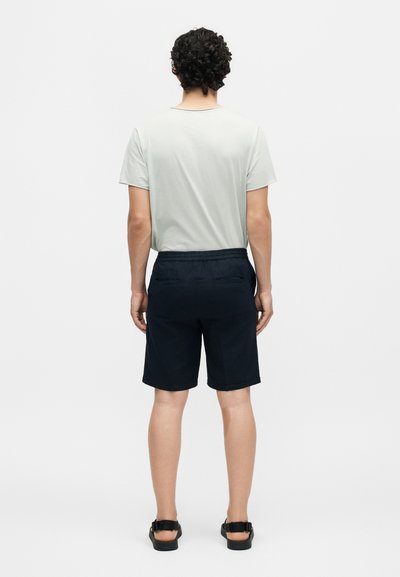 Άνδρας με σγουρά μαλλιά που φοράει λευκό πουκάμισο με κοντά μανίκια, σκούρα μπλε σορτς με πίσω τσέπες και μαύρα σανδάλια, στέκεται με την πλάτη προς τα έξω.