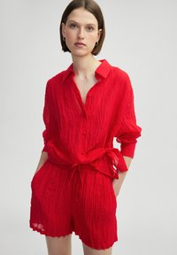 Camisa de tela roja y arrugada con botones en el frente, cuello y cintura ajustada. Combinada con pantalones cortos a juego que presentan una cintura elástica.
