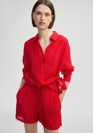 Camisa de tela roja y arrugada con botones en el frente, cuello y cintura ajustada. Combinada con pantalones cortos a juego que presentan una cintura elástica.
