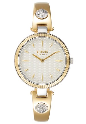 Montre Versus Versace en or et argent avec cadran blanc texturé, index dorés et accents tête de lion sur le bracelet.