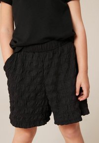 Pantalones cortos negros con textura, con tejido recogido, cinturilla elástica y un ajuste holgado. Cuenta con bolsillos y un dobladillo ondulado en la parte inferior.