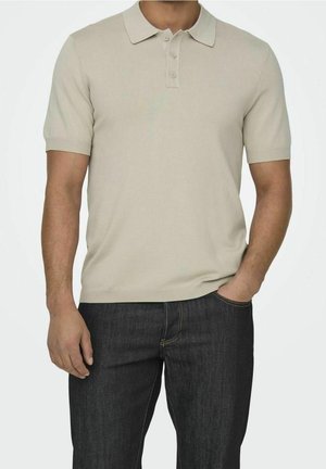 Man met een beige poloshirt met korte mouwen en donkerblauwe jeans, met één hand in de zak en een neutrale achtergrond.