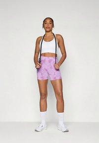 Reggiseno sportivo bianco, pantaloncini tie-dye viola e scarpe da ginnastica bianche. Modella tiene una corda per saltare, in piedi su uno sfondo neutro.