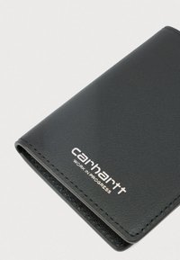 Μαύρο διπλωμένο δερμάτινο πορτοφόλι με το λογότυπο "Carhartt Work in Progress" τυπωμένο σε λευκό στη μπροστινή γωνία, σε λευκό φόντο.